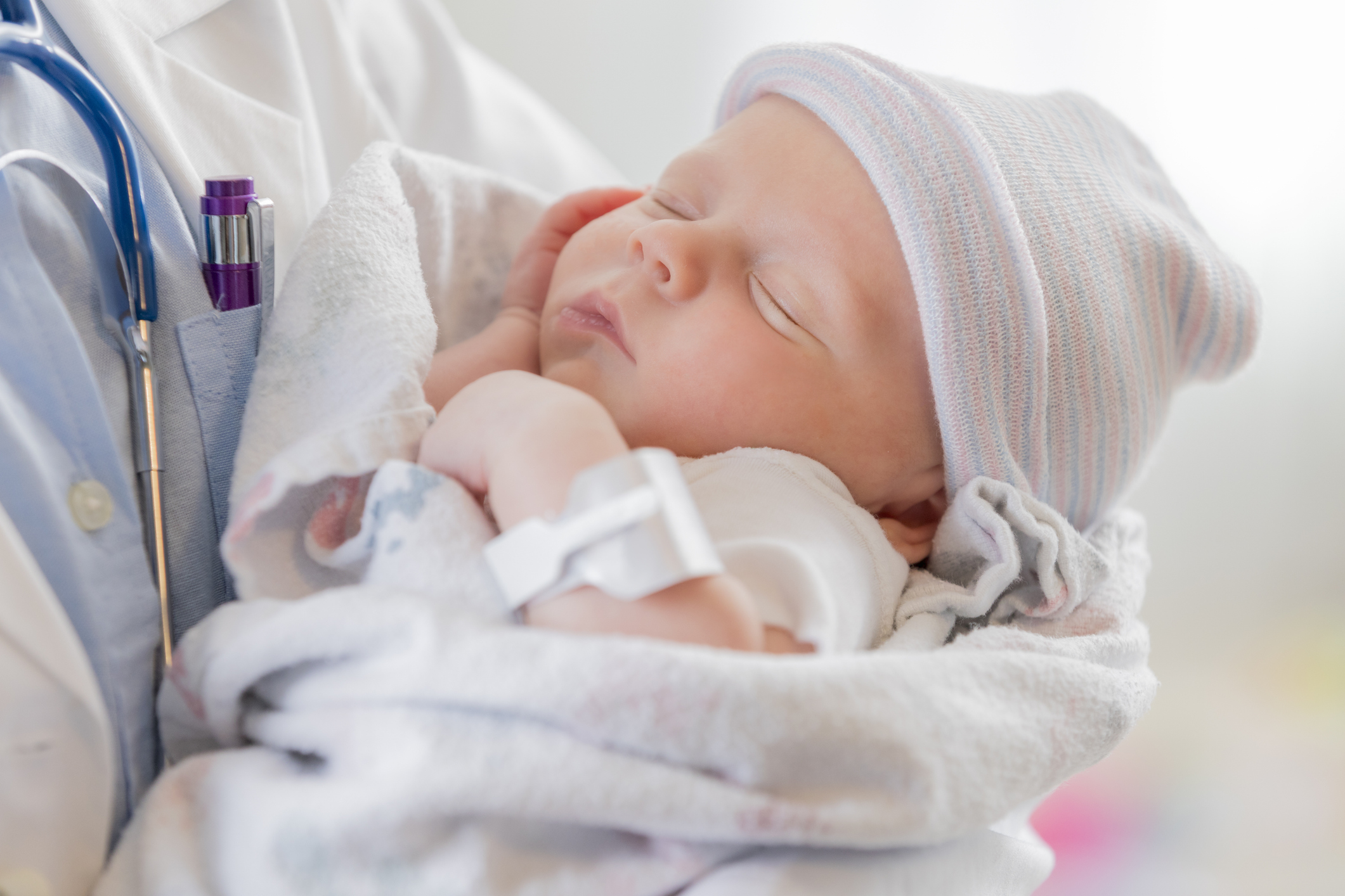 A Caucasian female infant is asleep in the arms of a pediatrician. The baby is swaddled in a blanket and is wearing a warm cap.